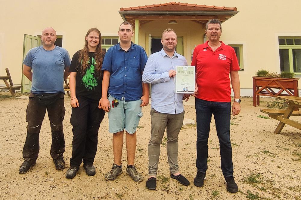 KJB Nickern erhält Preisgeld von der Town & Country Stiftung | vor dem Kinderbauernhaus stehen fünf Mitarbeiter des Kinderbauernhofes. Der Geschäftsführer (zweite von rechts) hält eine Urkunde in die Kamera. Rechts neben ihm steht ein Vertreter der Town & Country Stiftung